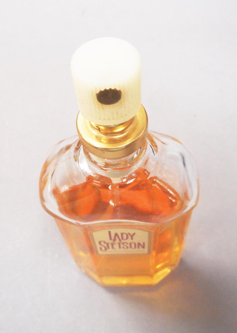 Puede incluir: Un frasco de perfume Lady Stetson vintage. El frasco de vidrio transparente tiene forma octogonal y contiene un l&iacute;quido naranja dorado. El frasco tiene un cuello dorado y una tapa color crema. La etiqueta dice "Lady Stetson".