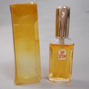 Op de afbeelding: Een heldere glazen parfumfles met een goudkleurige dop en het label "Lady Stetson" in het rood. De fles is gevuld met een goudkleurige vloeistof en staat naast een transparante, amberkleurige plastic doos.