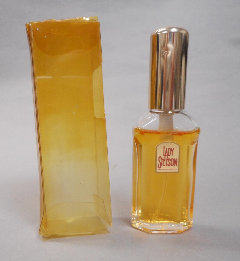 Op de afbeelding: Een heldere glazen parfumfles met een goudkleurige dop en het label "Lady Stetson" in het rood. De fles is gevuld met een goudkleurige vloeistof en staat naast een transparante, amberkleurige plastic doos.