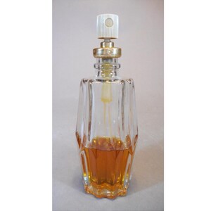 May include: A clear, faceted glass perfume bottle with a gold-colored collar and a white and gold atomizer. The bottle contains a golden-brown liquid. The bottle has a decorative, geometric design and a unique shape.