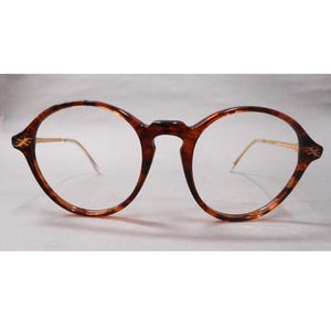 May include: Round eyeglasses with a tortoiseshell pattern frame. The glasses have gold-colored temples and decorative accents on the outer corners. The lenses are clear.