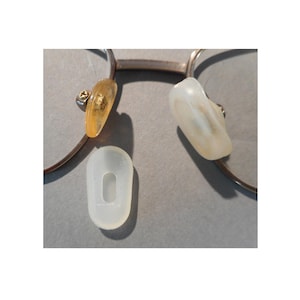 May include: Close-up of eyeglasses with detached nose pads. The metal frame is silver-toned, and the nose pads are a translucent yellow and white. One replacement pad is also visible, showing the oval shape and the hole for the nose bridge.
