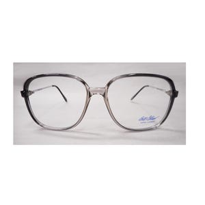 May include: A pair of eyeglasses with a clear frame and dark gray accents. The lenses are clear, and the temples are thin and silver. The glasses have a rectangular shape with rounded edges. The brand name is visible on the lower right lens.