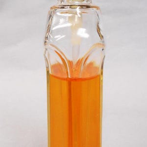 Puede incluir: Botella de perfume de vidrio transparente con cuello dorado y tap&oacute;n blanco. La botella est&aacute; llena de l&iacute;quido naranja. La botella tiene una forma &uacute;nica con base cuadrada y parte superior redondeada.