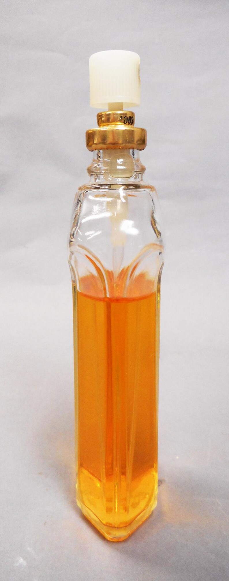 Puede incluir: Botella de perfume de vidrio transparente con cuello dorado y tap&oacute;n blanco. La botella est&aacute; llena de l&iacute;quido naranja. La botella tiene una forma &uacute;nica con base cuadrada y parte superior redondeada.