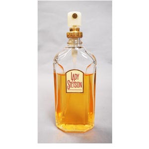 Puede incluir: Un frasco de perfume de vidrio transparente con una tapa dorada y una parte superior blanca. El frasco contiene un l&iacute;quido de color dorado y tiene una etiqueta que dice "LADY STETSON". El frasco tiene forma rectangular con bordes redondeados.