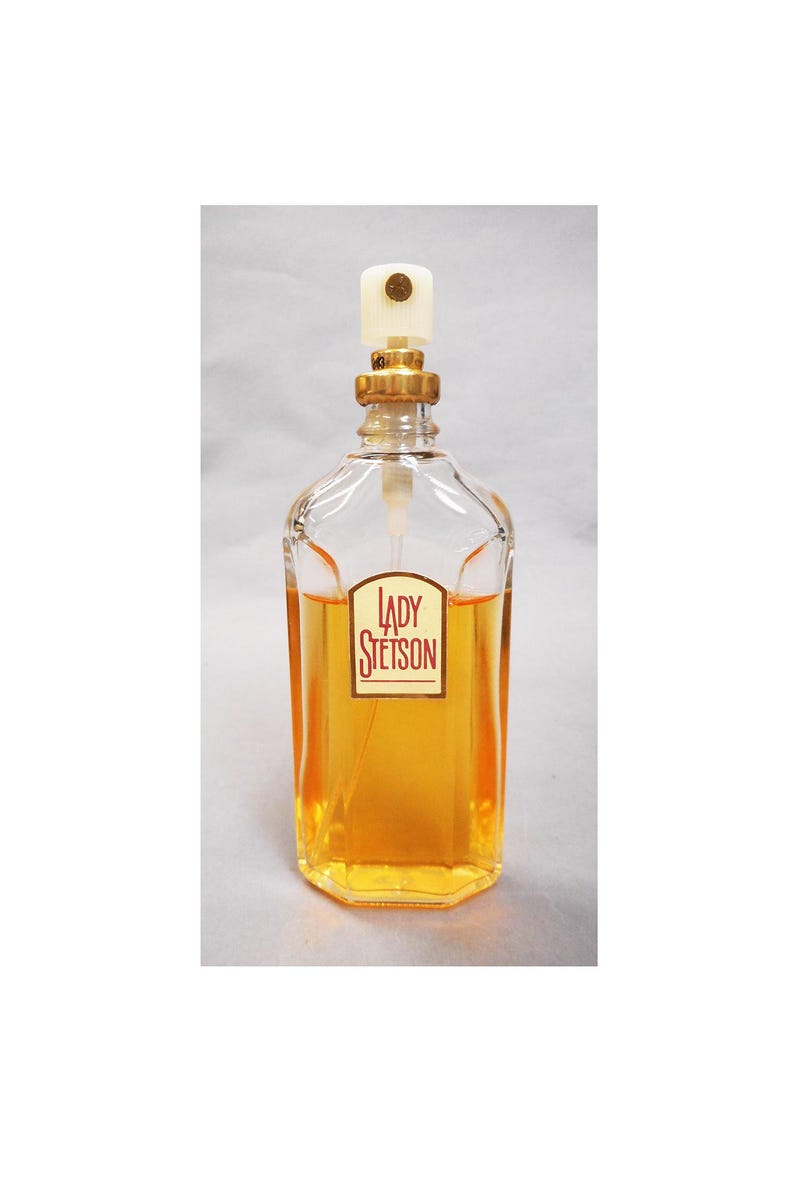 Puede incluir: Un frasco de perfume de vidrio transparente con una tapa dorada y una parte superior blanca. El frasco contiene un l&iacute;quido de color dorado y tiene una etiqueta que dice "LADY STETSON". El frasco tiene forma rectangular con bordes redondeados.
