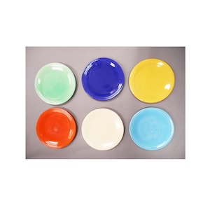 May include: A set of six round ceramic plates arranged in two rows of three. The plates feature a glossy finish and are colored in light green, dark blue, yellow, orange, off-white, and light blue, each with a concentric ring pattern.