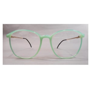 May include: Eyeglasses featuring a translucent mint green plastic frame with a rounded shape. The thin, metallic gold-toned temples connect to the frame front, ending in curved black plastic tips. The lenses are clear and transparent.