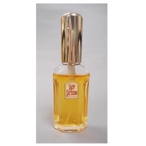 Op de afbeelding: Een heldere parfumfles met een goudkleurige dop en een label met de tekst "LADY Stetson". De fles bevat een gele vloeistof. De fles heeft een rechthoekige vorm met een goudkleurige dop.