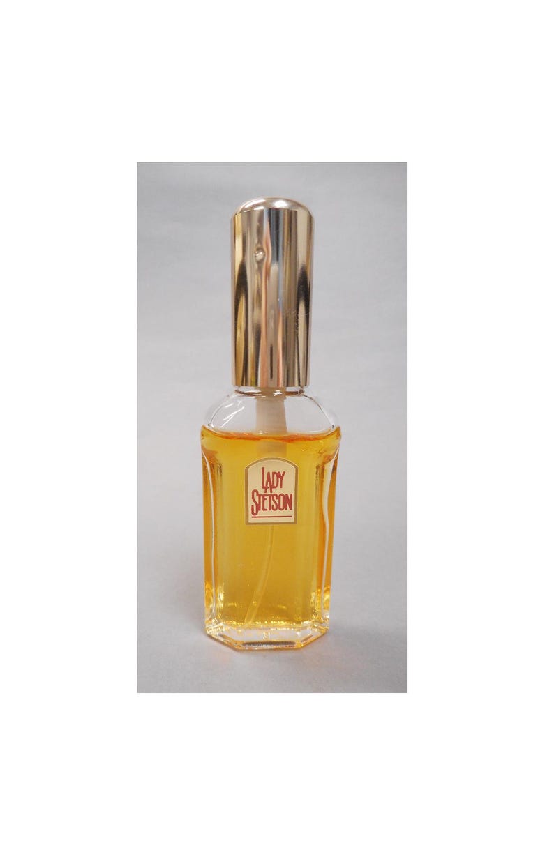 Op de afbeelding: Een heldere parfumfles met een goudkleurige dop en een label met de tekst "LADY Stetson". De fles bevat een gele vloeistof. De fles heeft een rechthoekige vorm met een goudkleurige dop.