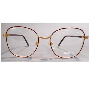 May include: Eyeglasses with a gold bridge and temples, and a burgundy-colored frame. The lenses are clear, and the brand name "Zimco" is printed on the lower right lens.