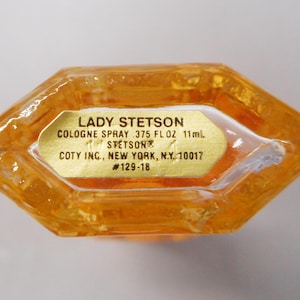 Op de afbeelding: Een vintage Lady Stetson eau de cologne sprayfles. De amberkleurige glazen fles heeft een unieke achthoekige vorm. Een gouden etiket toont de productinformatie, inclusief het volume van 11 ml. Het etiket bevat ook de gegevens van de fabrikant.