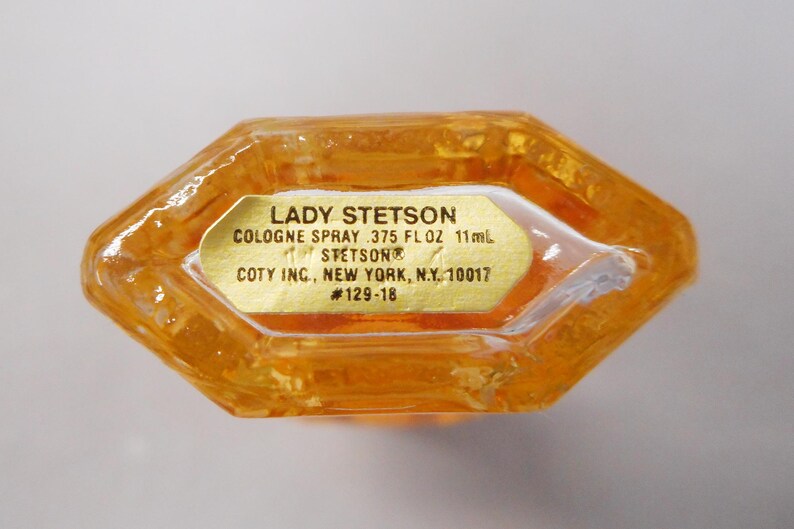 Op de afbeelding: Een vintage Lady Stetson eau de cologne sprayfles. De amberkleurige glazen fles heeft een unieke achthoekige vorm. Een gouden etiket toont de productinformatie, inclusief het volume van 11 ml. Het etiket bevat ook de gegevens van de fabrikant.
