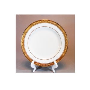 May include: A round white porcelain plate featuring a wide decorative border with a gold and brown checkered pattern. The plate has a thin gold rim and a thin gold inner circle accent, resting on a white plastic display stand for upright viewing.