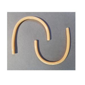 May include: Two curved, light beige tubes are arranged in a semi-circle shape against a gray background. The tubes appear to be made of a smooth, possibly plastic material. The ends of the tubes are cut straight, and the overall design is simple and minimalist.
