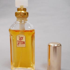 Op de afbeelding: Een heldere parfumfles van glas met een goudkleurige dop en accenten. De fles bevat een gele vloeistof en heeft een label met de tekst "Lady Stetson". De dop staat ernaast.