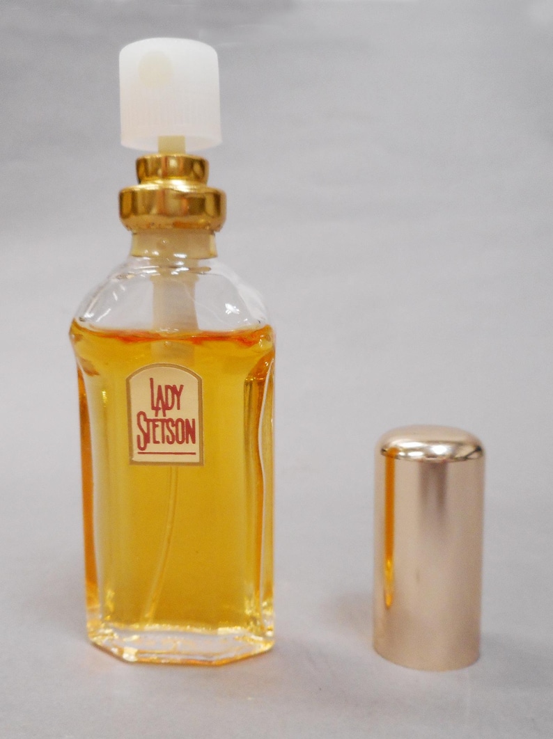 Op de afbeelding: Een heldere parfumfles van glas met een goudkleurige dop en accenten. De fles bevat een gele vloeistof en heeft een label met de tekst "Lady Stetson". De dop staat ernaast.