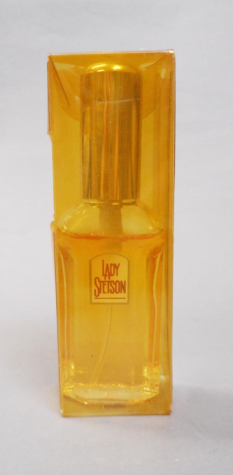 Op de afbeelding: Een fles Lady Stetson parfum in een heldere, geel getinte rechthoekige fles, verpakt in een bijpassende gele plastic doos. Het etiket op de fles luidt "Lady Stetson".