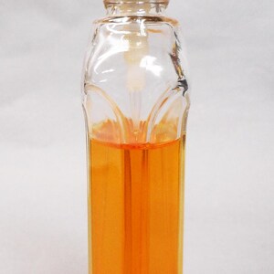 Puede incluir: Un frasco de perfume de vidrio transparente con tap&oacute;n blanco y detalles dorados. La botella est&aacute; llena de un l&iacute;quido naranja. La botella tiene forma rectangular con un dise&ntilde;o decorativo en la parte delantera.