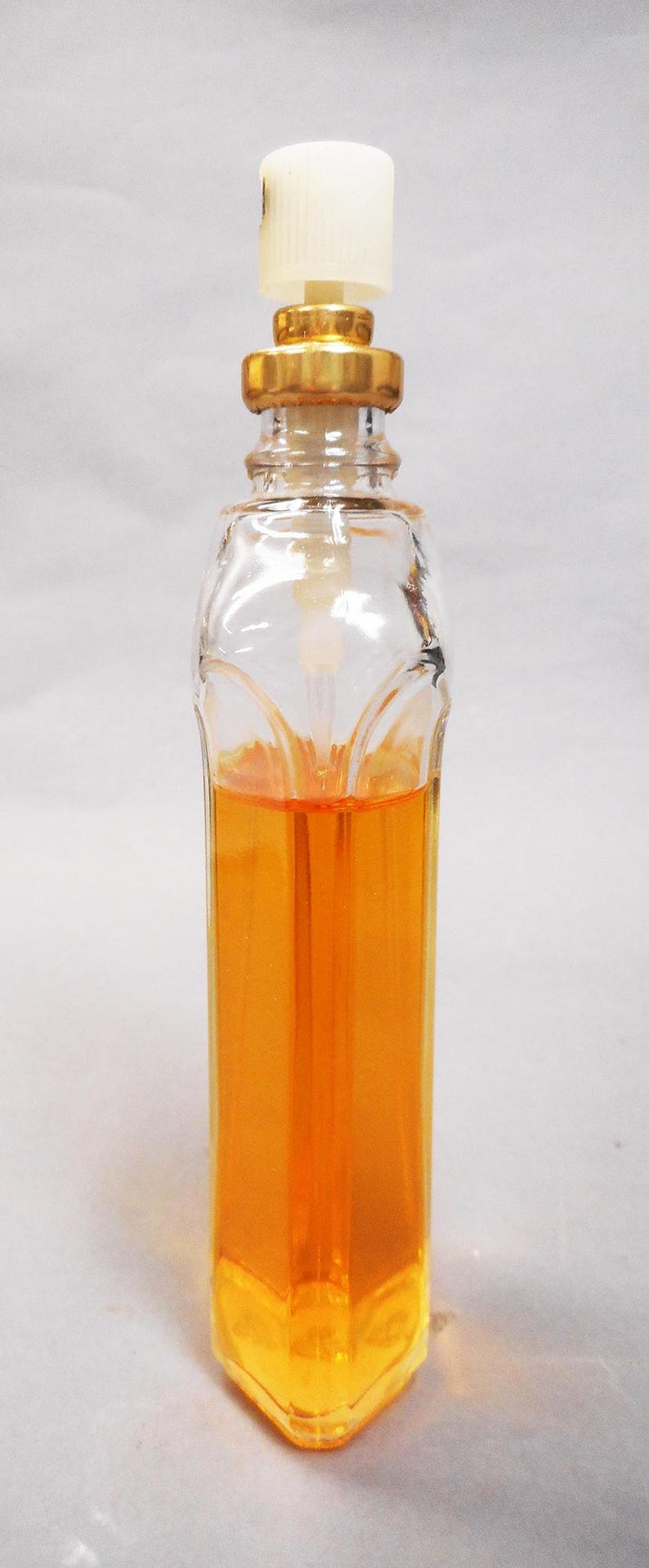 Puede incluir: Un frasco de perfume de vidrio transparente con tap&oacute;n blanco y detalles dorados. La botella est&aacute; llena de un l&iacute;quido naranja. La botella tiene forma rectangular con un dise&ntilde;o decorativo en la parte delantera.