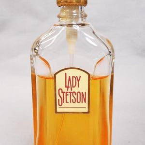Puede incluir: Una botella de vidrio transparente de perfume Lady Stetson. La botella tiene una tapa dorada y un pulverizador blanco. El perfume en su interior es de color &aacute;mbar dorado. La etiqueta frontal dice "LADY STETSON" en un dise&ntilde;o rojo y blanco.