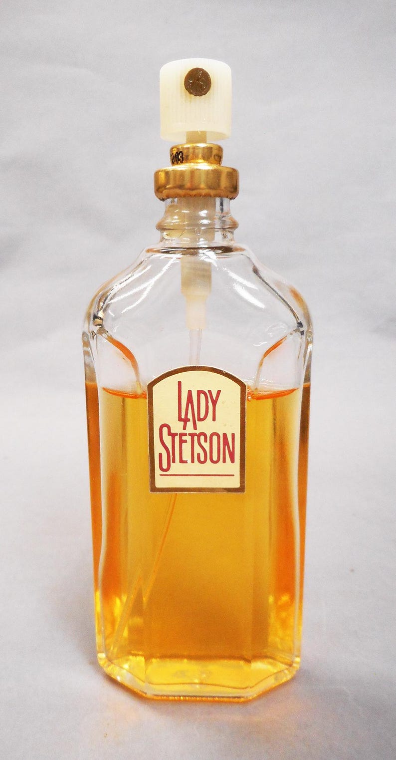 Puede incluir: Una botella de vidrio transparente de perfume Lady Stetson. La botella tiene una tapa dorada y un pulverizador blanco. El perfume en su interior es de color &aacute;mbar dorado. La etiqueta frontal dice "LADY STETSON" en un dise&ntilde;o rojo y blanco.