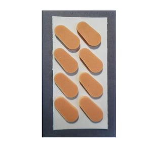 May include: A sheet of eight small, oblong, peach-colored foam pads arranged in two columns of four. The pads are on a white rectangular backing, likely for adhesive application. The background is a neutral gray.