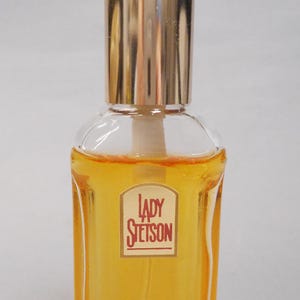 Op de afbeelding: Een heldere glazen parfumfles met een goudkleurige dop. De fles bevat een gele vloeistof en heeft een rechthoekig etiket met de woorden "LADY STETSON" in rode letters.