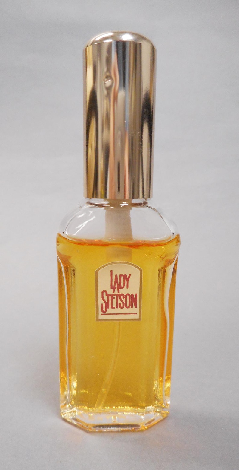Op de afbeelding: Een heldere glazen parfumfles met een goudkleurige dop. De fles bevat een gele vloeistof en heeft een rechthoekig etiket met de woorden "LADY STETSON" in rode letters.