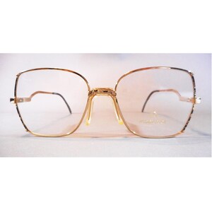 May include: Gold-toned eyeglasses with a square frame design. The glasses have a thin gold frame with a double bridge and black temples. The lenses are clear, and the brand name is visible on the right lens.