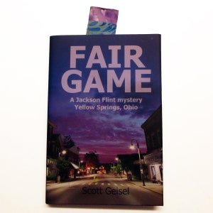 以下が含まれることがあります： 「フェアゲーム」というタイトルのついた本で、紫と青のしおりがついています。本の表紙は、紫とピンクの空の下、小さな町の通りの夜のシーンが描かれています。表紙のテキストには、「A Jackson Flint mystery Yellow Springs, Ohio」と「Scott Geisel」と書いてあります。