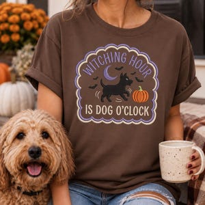 May include: A brown short-sleeved t-shirt featuring a graphic print of a black dog, a pumpkin, a crescent moon, and bats. The text reads "WITCHING HOUR IS DOG O'CLOCK" inside a scalloped purple border. The design is centered on the chest area.