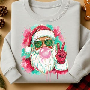 May include: A light gray sweatshirt featuring a graphic of Santa Claus blowing a bubblegum bubble. Santa is wearing sunglasses with snowflake lenses and giving a peace sign. The design has pink and teal watercolor accents.