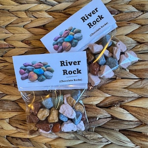 Pode incluir: Duas sacolas de pedras de chocolate, etiquetadas como "River Rock (Chocolate Rocks)". As sacolas estão sobre uma superfície de vime trançado.