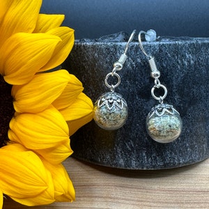 May include: A pair of silver dangle earrings with clear glass orbs filled with green and brown dried herbs. The earrings are hanging from a gray stone surface.
