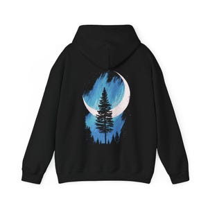 May include: Black hoodie featuring a white crescent moon and a silhouetted pine tree design. The artwork includes blue and white brushstrokes, and a forest silhouette at the bottom. A cozy, casual garment.
