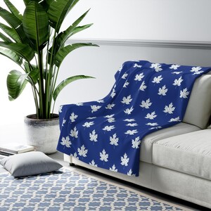 May include: A blue throw blanket with a white maple leaf pattern is draped over a white sofa. The blanket is made of a soft material and is a decorative home accessory. The blanket is a square or rectangular shape.
