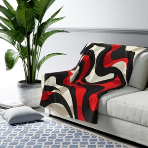 May include: A plush throw blanket featuring a bold, abstract pattern of flowing, interconnected waves in solid black, vibrant red, and soft cream colors. The fabric has a smooth texture and is draped over the corner of a neutral-colored sofa.