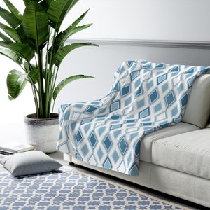 Peut inclure: Une couverture blanche avec un motif géométrique de diamants bleus et blancs. La couverture est posée sur un canapé gris clair.