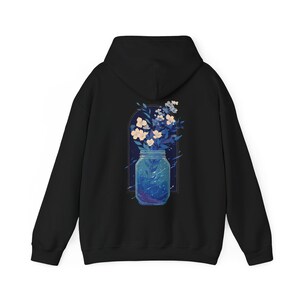 May include: Black hoodie featuring a vibrant illustration of a jar filled with a starry night design and flowers. The jar is framed by an arch, with white and blue flowers spilling out. The hoodie has a hood and long sleeves.