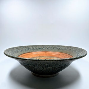 May include: A large, wide, ceramic serving bowl with a speckled green glaze and a brown rim. The bowl has a slightly raised lip around the edge.