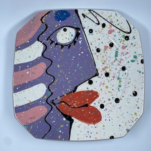 May include: A square ceramic plate with a colorful abstract design of a face. The face is painted in shades of purple, pink, white, and black. The background is white with black and colored dots.