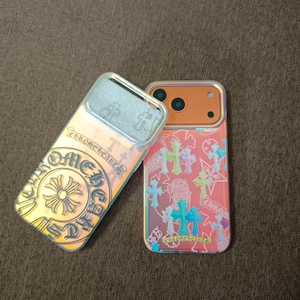Puede incluir: Dos fundas iridiscentes para teléfono. Una funda transparente con el texto "Chrome Hearts" y un diseño floral. La otra es roja con un patrón de cruces de varios colores. Ambas fundas tienen un acabado metálico.