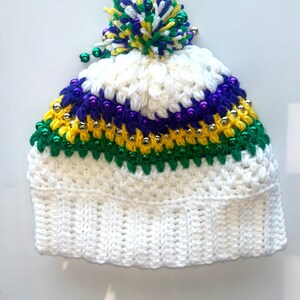 May include: White crocheted beanie with a green, yellow, and purple striped band and a green, yellow, and white pom pom. The beanie is decorated with gold and purple beads.