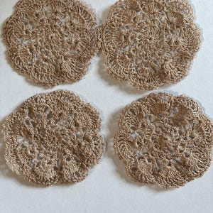 May include: Four beige, crocheted coasters arranged in a square. Each coaster has a floral design with a scalloped edge. The coasters are made of a light brown yarn and are placed on a white surface.