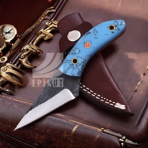 May include: A small, blue and black knife with a leather sheath. The knife has a blue handle with a marbled pattern and gold accents. The blade is black and has a slight curve.