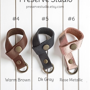 May include: Three leather keychains in warm brown, dark gray, and rose metallic. Each keychain has a metal button and a metal ring. The text "Preserve Studio" and the Etsy website address are at the top.