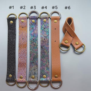 May include: A collection of six leather straps with metal D-rings and rivets. The straps vary in color and texture, including black, pink with gold spots, iridescent, and tan. Each strap is labeled with a number from #1 to #6.