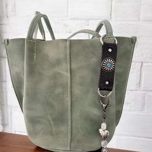 May include: A sage green leather tote bag with a black leather strap and a silver charm with a turquoise stone. The bag has a top handle and a shoulder strap.